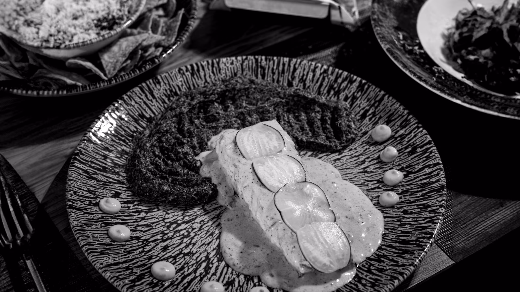 A plated dish in the foreground on a dinner table, in black and white