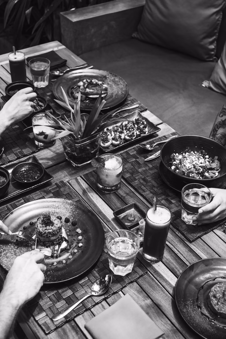 Table de restaurant garnie de plats et de verres, mains autour, en noir et blanc