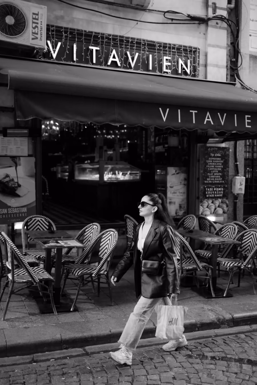 Femme marchant devant la terrasse d'un café de quartier, en noir et blanc